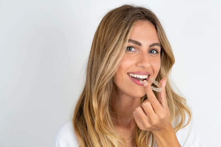 Invisalign - young blonde woman wearing white shirt over white studio background holding an invisible aligner ready to use it.