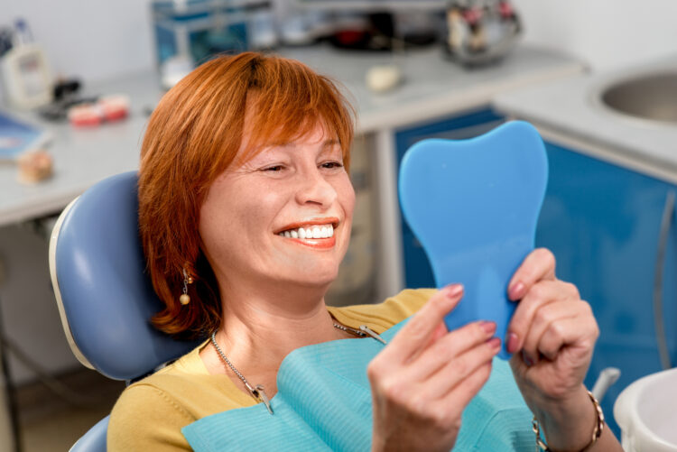 Dentures - Smiling senior woman with new dental implants sitting in the dental office and looking at the mirror